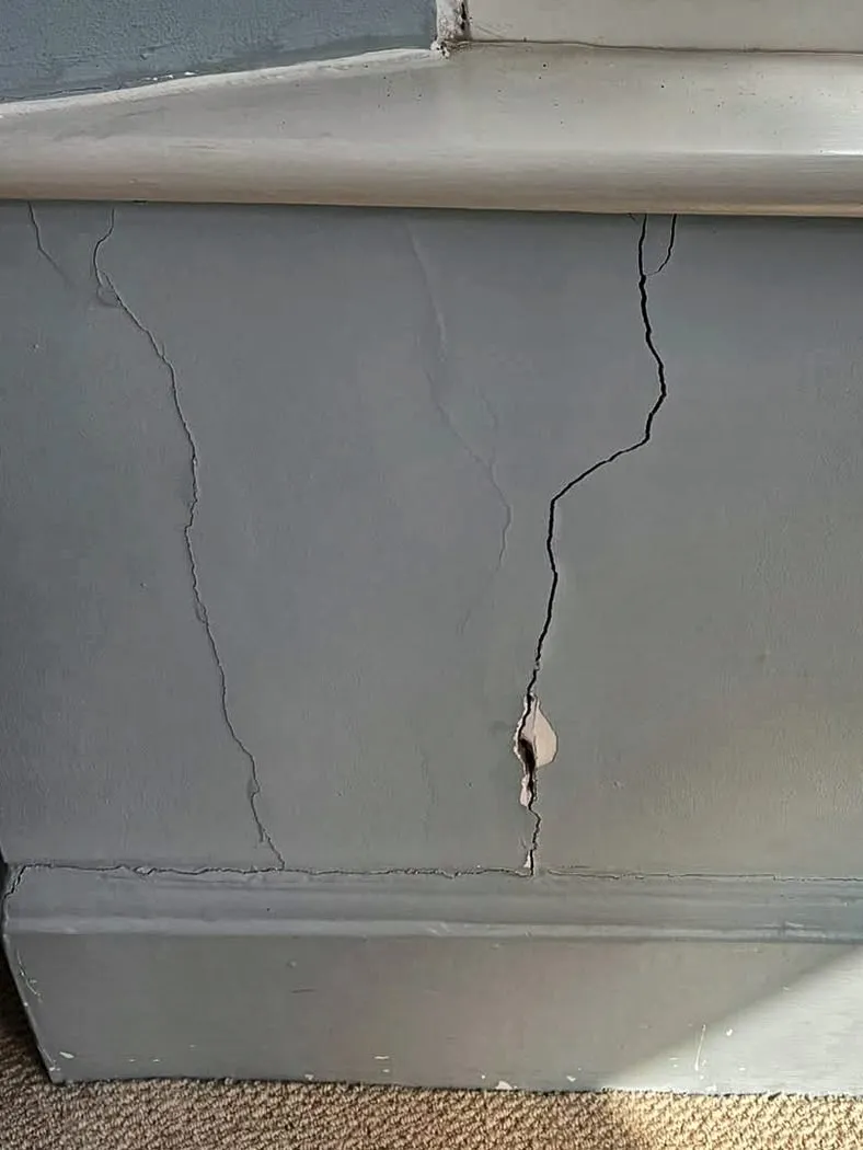 Two cracks in the plaster under a window board running down to the skirting, with a flake of plaster broken away