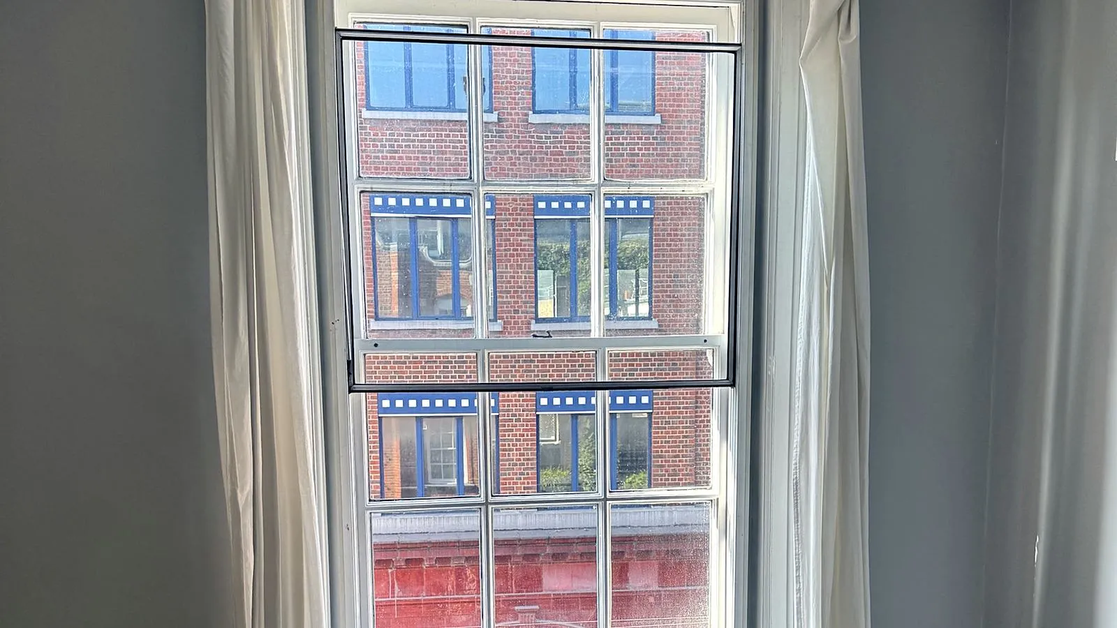 Tall single-glazed timber sash window with white curtains in a period room, with a slim panel fitted on the inside