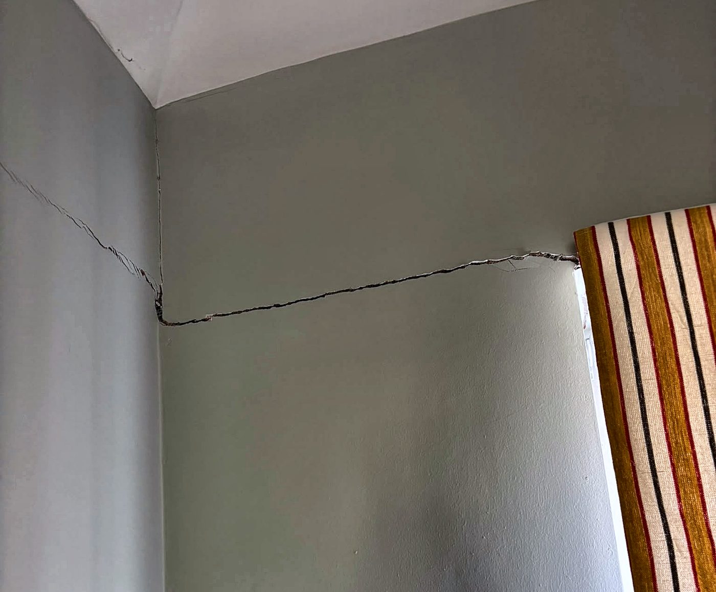 A wide crack running from the top corner of a window across the wall to the corner of the room, then continuing up the adjoining wall towards the ceiling