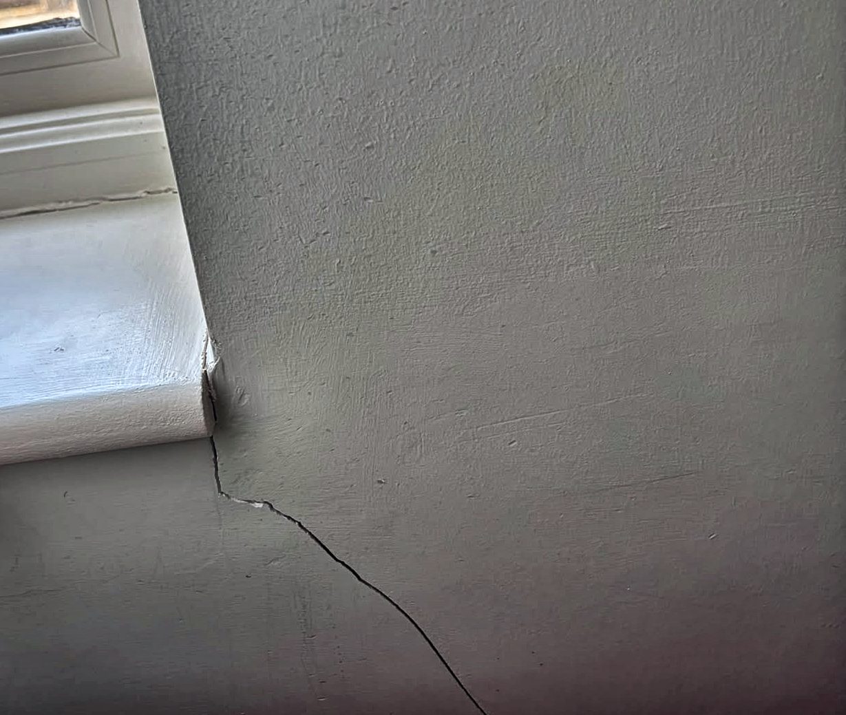 A diagonal crack running from the end of a painted window board down across the plaster towards the floor