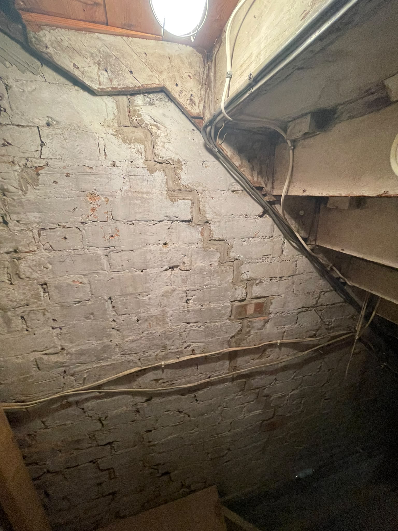 A stepped diagonal crack running down through the mortar joints of a painted brick wall under a staircase, with pale lines of earlier repair mortar along the crack