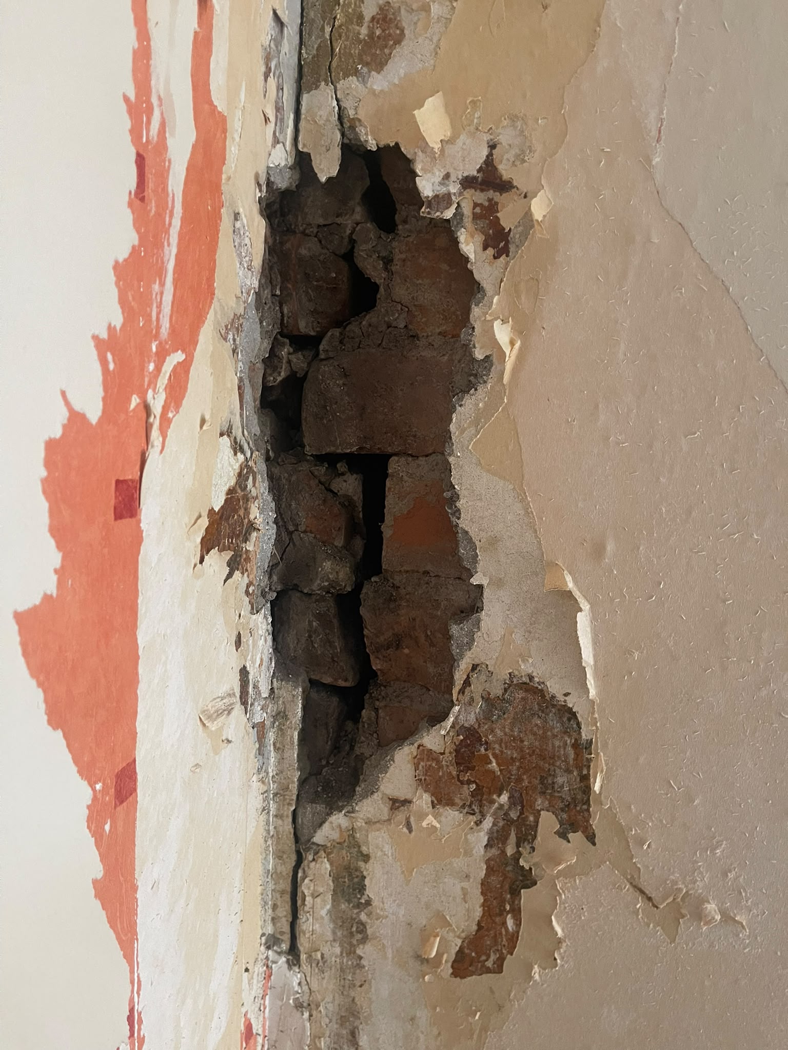 Wallpaper and plaster broken away from a vertical crack in an internal wall, exposing brickwork with an open vertical gap running through it